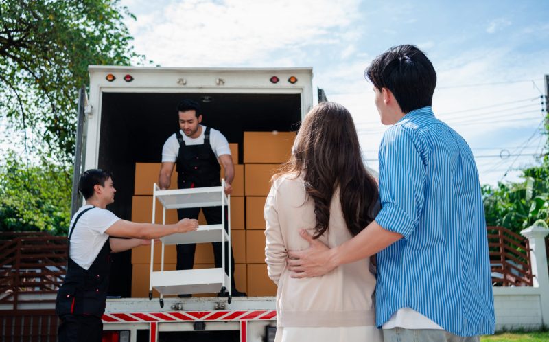 Asian Couple check while unloading boxes and furniture from a pickup truck to a new house with service cargo two men movers worker in uniform lifting boxes. concept of Home moving and delivery.